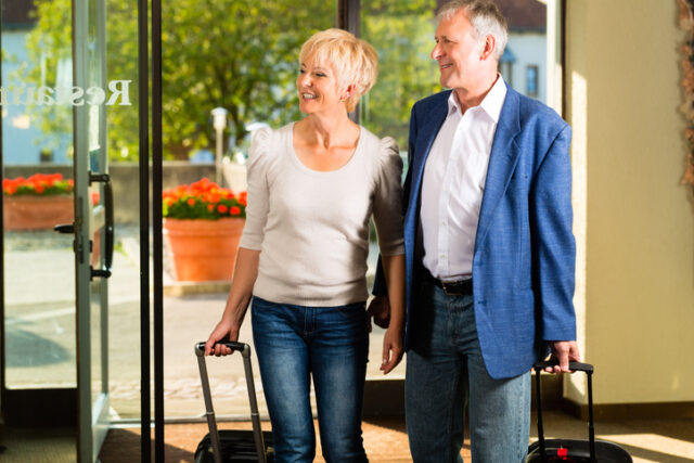 A senior man and woman traveling with rolling luggage, entering a building for a family visit.